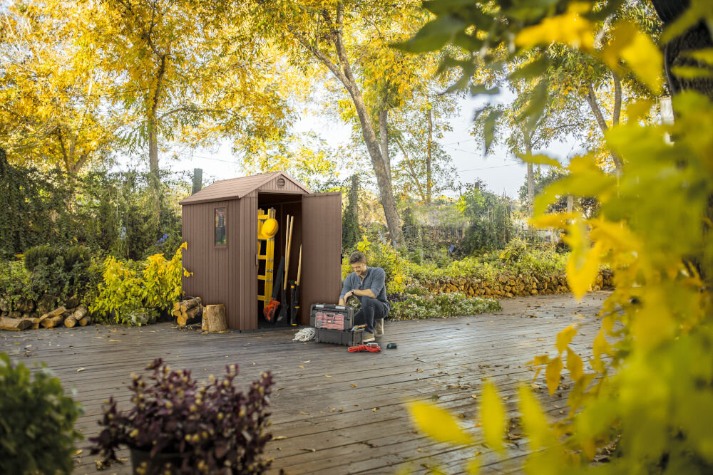 Na drewnianym tarasie stoi brązowy domek narzędziowy z otwartymi drzwiami, w którym widać narzędzia ogrodowe. Obok domku siedzi osoba z narzędziami, otoczona zielenią i żółtymi liśćmi drzew.