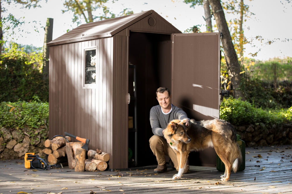 Brązowy domek narzędziowy z otwartymi drzwiami stoi na drewnianym tarasie, obok którego leżą kawałki drewna i narzędzia. Przed domkiem znajduje się osoba z psem.
