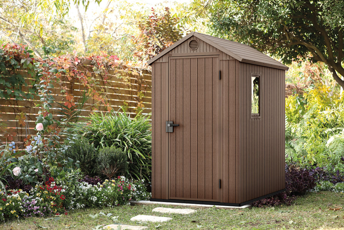 Brązowy domek narzędziowy z drzwiami i oknem, otoczony bujną roślinnością i kwiatami. W tle widoczny jest drewniany płot i drzewa.