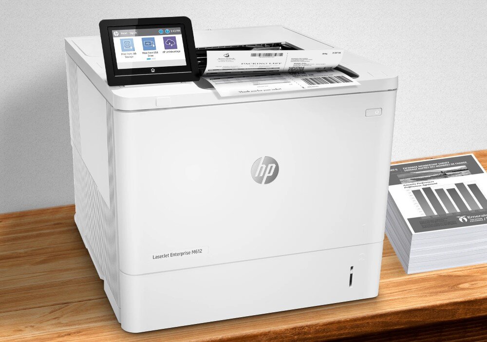 Drukarka HP LaserJet Enterprise M612dn Automatyczny druk dwustronny Drukarka umieszczona na drewnianym blacie drukuje etykiety, obok leży stos raportów, szybkość, prędkość druku, druk dwustronny