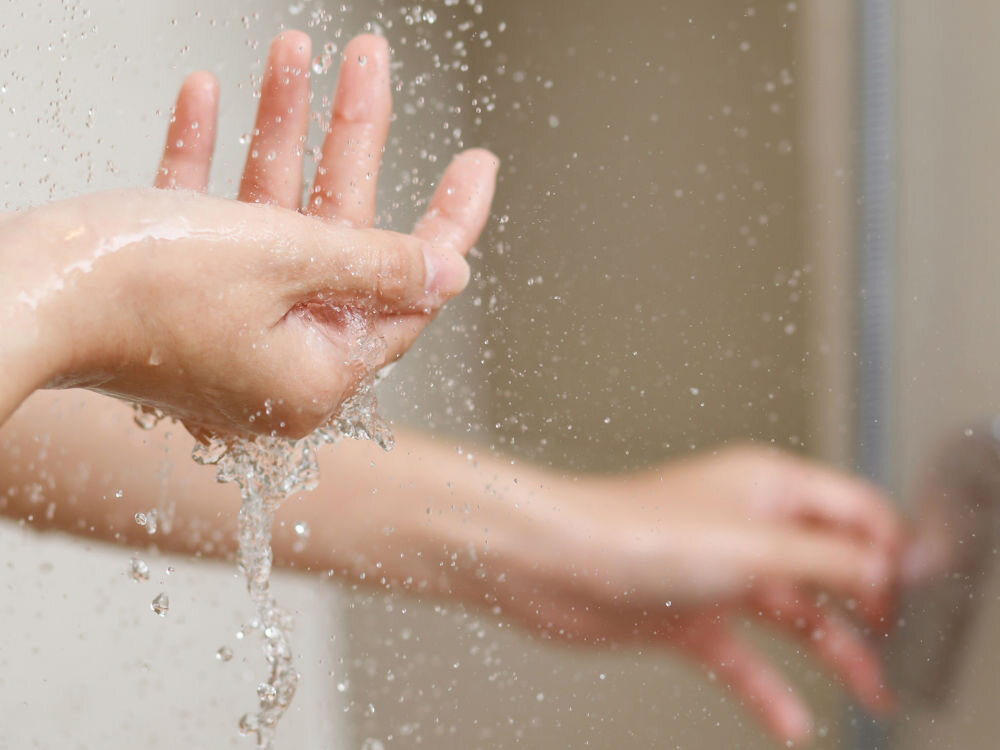 Na zdjęciu widać ręce pod strumieniem wody, z kroplami wody rozpryskującymi się wokół. Tło jest rozmyte, co sugeruje, że scena ma miejsce w łazience lub pod prysznicem.
