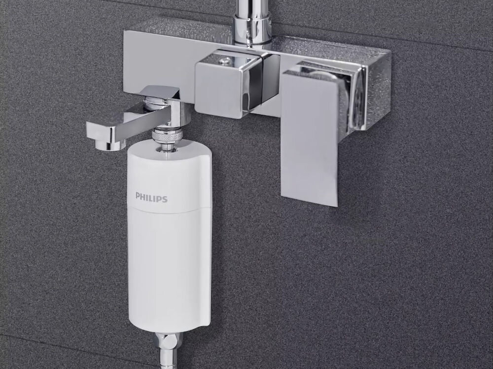 Na obrazie widoczny jest biały filtr prysznicowy z napisem \'PHILIPS\', zamontowany na metalowej armaturze łazienkowej. Tło stanowi ciemna, gładka powierzchnia ściany.