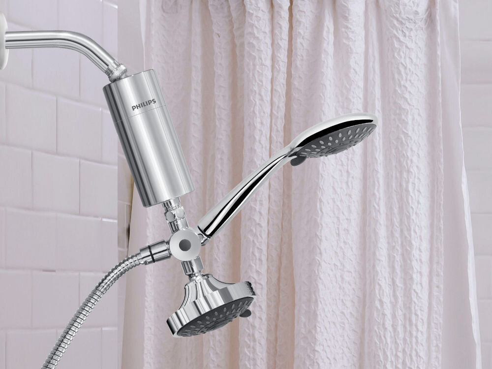 Na obrazie widoczny jest srebrny filtr prysznicowy z logo PHILIPS, zamontowany na prysznicu z metalowym wężem. W tle znajduje się biała zasłona prysznicowa i kafelki.