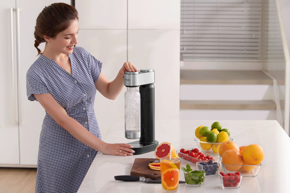 Saturator PHILIPS ADD4902BK/10 GoZero Uśmiechnięta kobieta obsługująca czarny saturator Philips na blacie kuchennym, przygotowując wodę gazowaną obok miseczek ze świeżymi owocami.