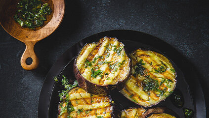 Na czarnym talerzu znajdują się grillowane plastry bakłażana posypane ziołami. Obok na drewnianej łyżce widoczna jest zielona mieszanka ziół.