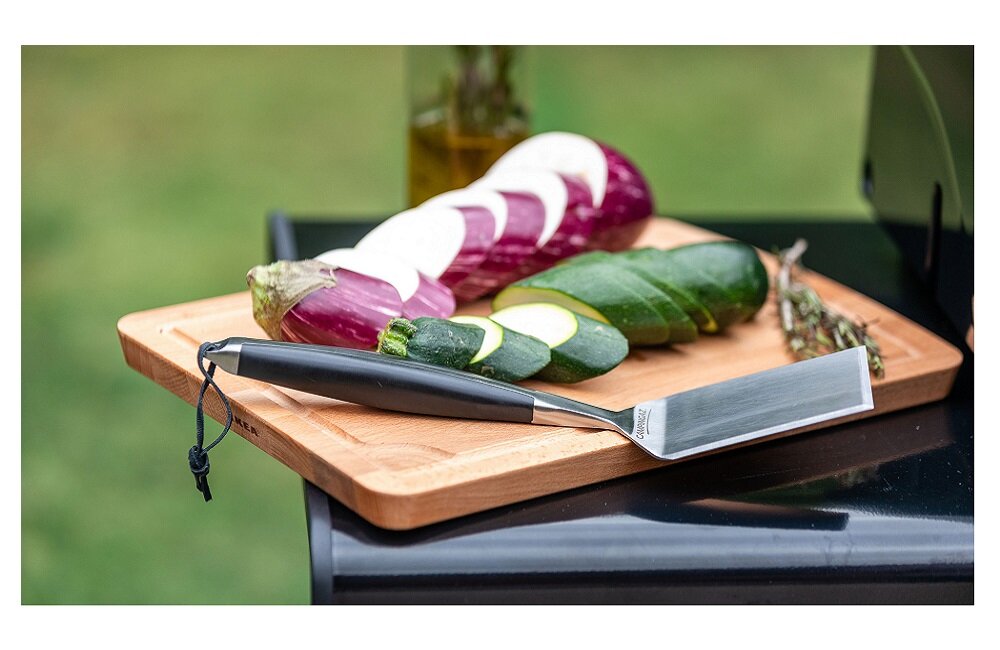 Na drewnianej desce do krojenia znajdują się pokrojone warzywa: bakłażan i cukinia, obok leży nóż z czarną rączką. W tle widoczna jest butelka z oliwą i gałązka ziół.