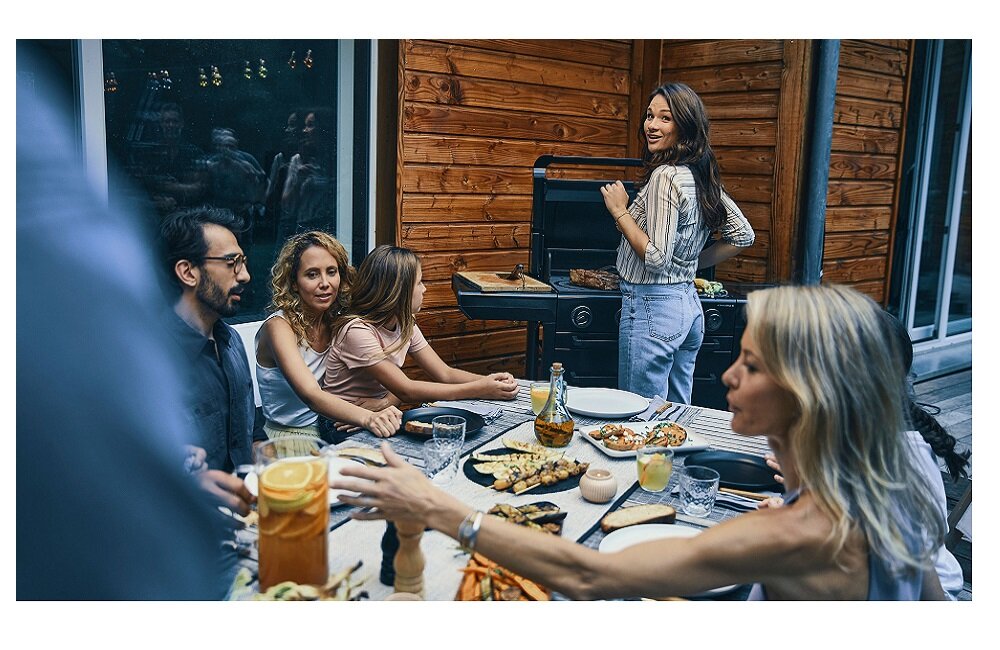 Grupa osób siedzi przy stole na zewnątrz, na którym znajdują się różne potrawy i napoje. W tle widoczny jest grill gazowy, przy którym stoi kobieta.
