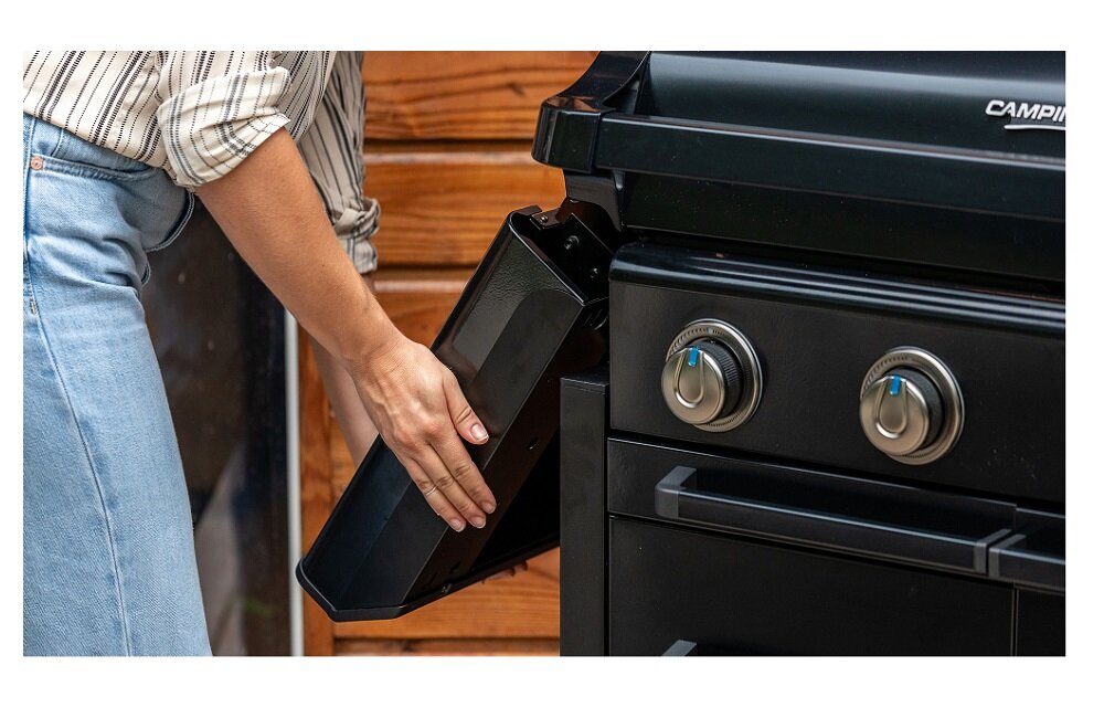Osoba wyciąga czarną tacę z grilla gazowego, na którym widoczne są dwa pokrętła i logo \'CAMPINGAZ\'.
