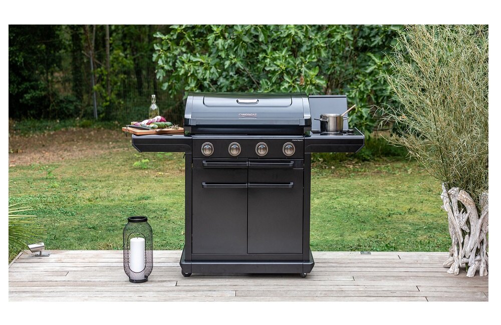 Na drewnianym tarasie stoi czarny grill gazowy z czterema pokrętłami i zamkniętą pokrywą. Obok grilla znajduje się butelka z napojem i talerz z jedzeniem, a w tle widoczna jest zieleń ogrodu.