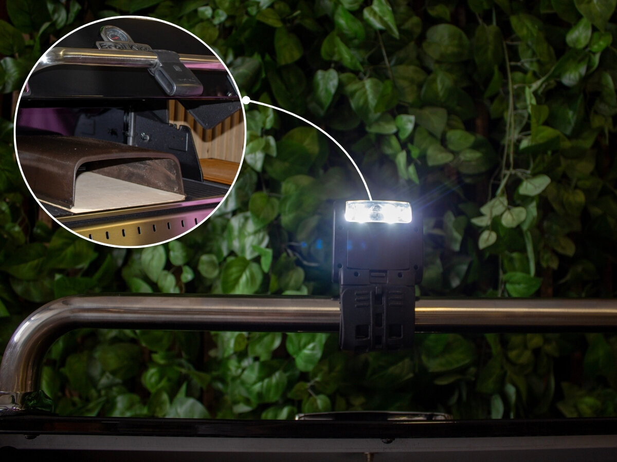 Latarka do rusztu BROIL KING 60934, komfort, grill, zmrok, światło, Broil King LED Grill Light zamontowany na rurze grilla Broil King oświetla obszar obok, z zaznaczonym uchwytem na akcesoria i zielenią w tle.