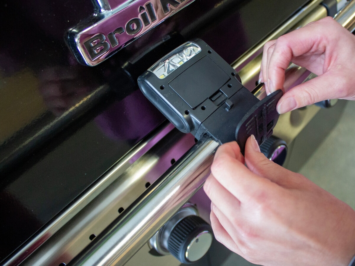 Latarka do rusztu BROIL KING 60934, montaż, pasek, uchwyt, stabilność, Dwie ręce montują czarną lampę LED Broil King na boku grilla gazowego Broil King obok pokręteł sterujących.