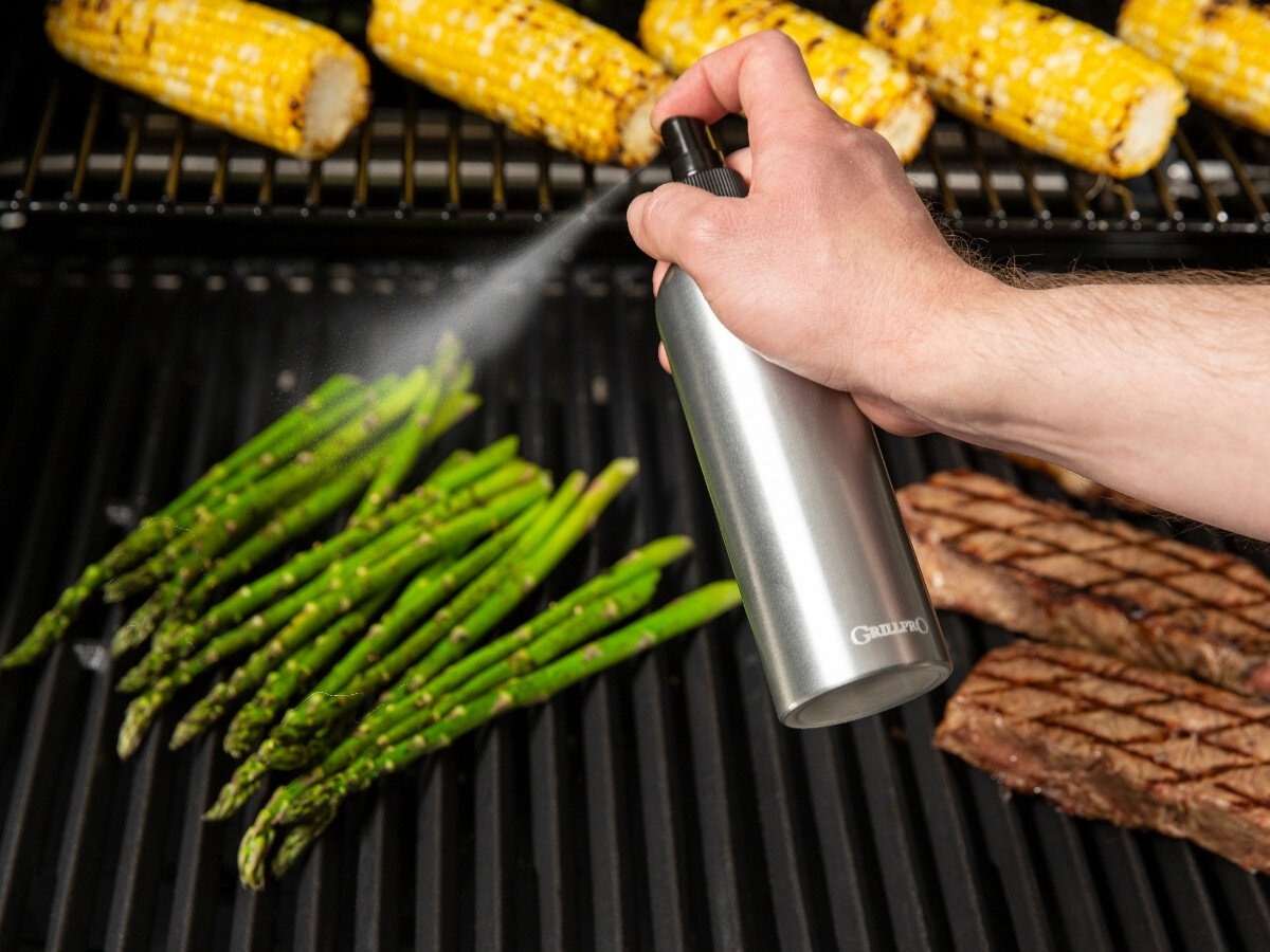 Rozpylacz oleju BROIL KING Grill Pro 50945, mgiełka oleju, równomierne rozpylenie, grill, precyzja, Ręka spryskuje olejem ze stalowej butelki szparagi leżące na ruszcie grilla, w tle widać grillowaną kukurydzę i steki.