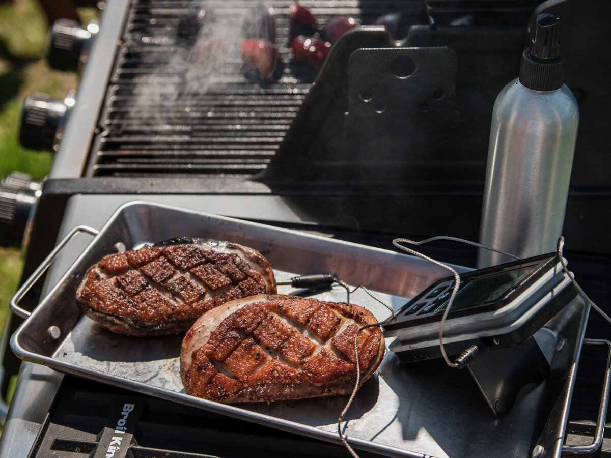 Rozpylacz oleju BROIL KING Grill Pro 50945, natłuszczanie potraw, soczyste mięso, chrupiące warzywa, grillowanie,Zbliżenie na tacę z soczystymi, przyrumienionymi stekami i termometrem grillowym na półce grilla, obok stoi metalowa butelka ze spryskiwaczem do oleju, w tle rozgrzany ruszt z warzywami.