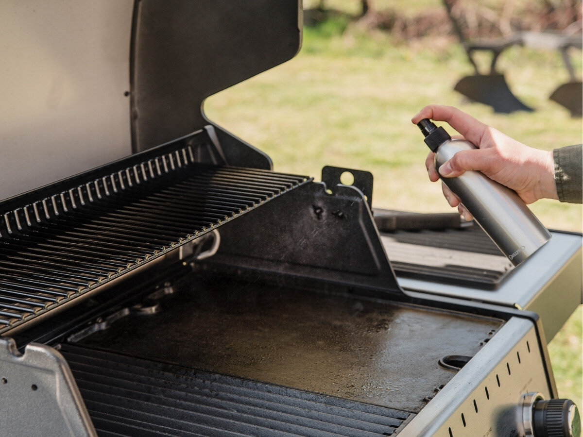 Rozpylacz oleju BROIL KING Grill Pro 50945, ruszt, płyta żeliwna, równomierne pokrycie, przygotowanie, Osoba spryskuje olejem czystą płytę i ruszt grilla gazowego za pomocą metalowej butelki ze spryskiwaczem na ogrodzie.