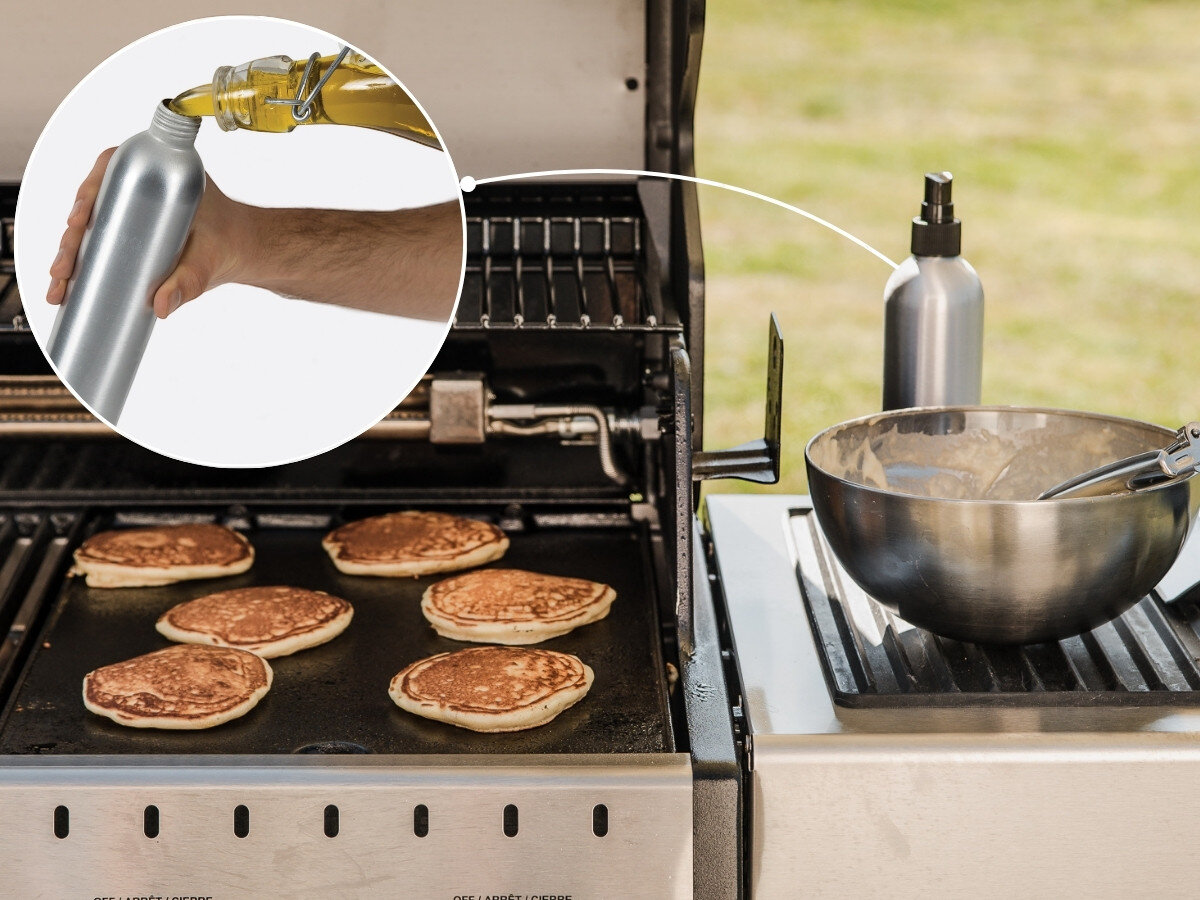Rozpylacz oleju BROIL KING Grill Pro 50945, zbiornik, 350 ml, zapas oleju, praktyczność, Metalowa butelka ze spryskiwaczem stojąca przy misce z ciastem na płycie grillowej, w tle naleśniki smażące się na rozgrzanej płycie grilla gazowego na świeżym powietrzu.