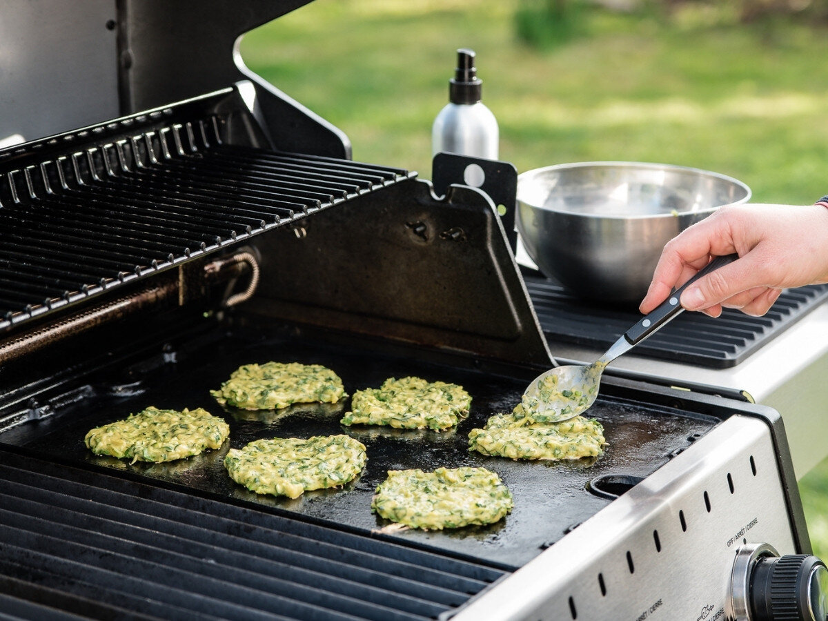 Rozpylacz oleju BROIL KING Grill Pro 50945, bez aerozolu, bezpieczeństwo, ekologia, wielokrotne użycie, Ręka nakłada łyżką placuszki z warzyw na płytę grilla gazowego, obok stoi metalowy spryskiwacz do oleju i miska z masą, w tle ogród.