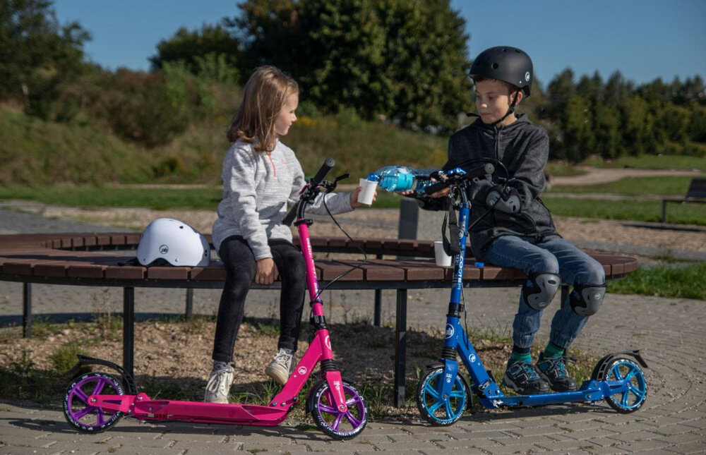 Hulajnoga PB Damper Różowy park na ławce w parku siedzi dwójka dzieci przed dziećmi stoją hulajnogi w tle niebo drzewa trawnik pasja hobby komfortowa jazda mobilność