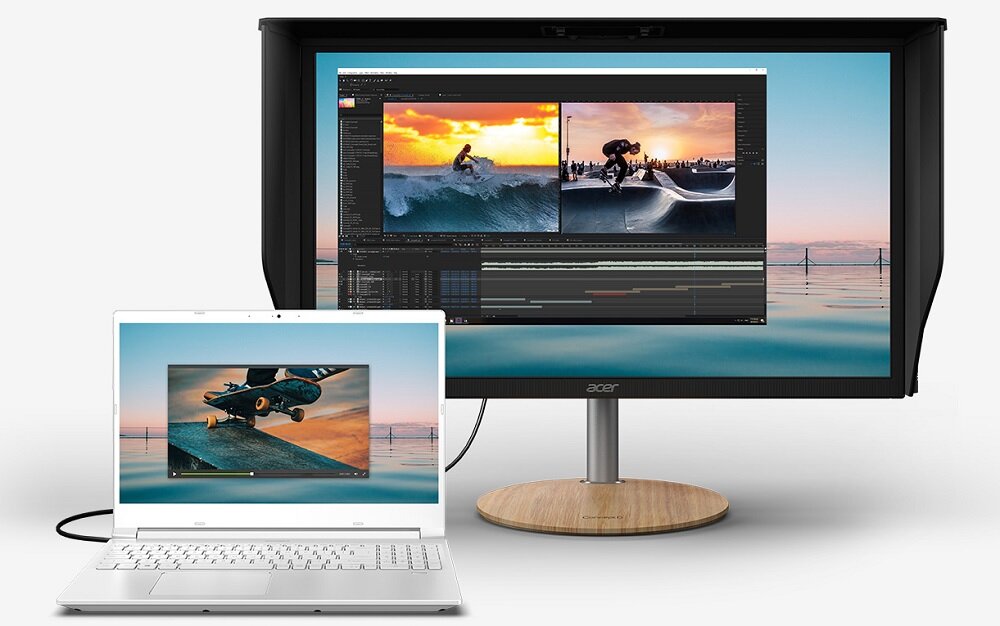 Na obrazie widoczny jest biały laptop z wyświetlonym zdjęciem skatera oraz duży monitor z dwoma zdjęciami surferów i oprogramowaniem do edycji wideo. Monitor stoi na drewnianej podstawce, a w tle widać spokojne morze.