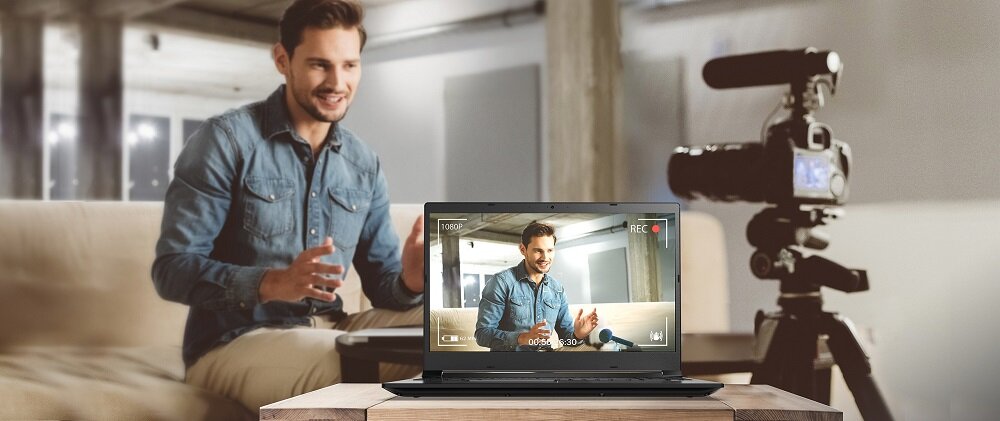 Na stole stoi laptop, na ekranie którego widać mężczyznę w niebieskiej koszuli, siedzącego na kanapie. Obok laptopa znajduje się kamera na statywie, a w tle widoczny jest ten sam mężczyzna.