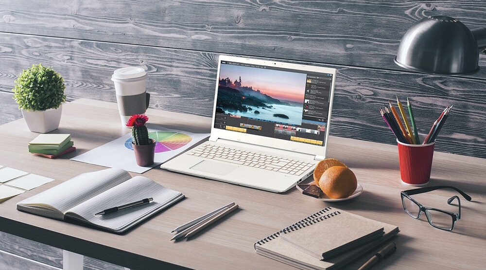 Na biurku znajduje się otwarty laptop z wyświetlonym programem do edycji wideo. Obok laptopa leżą notatnik, długopis, okulary, filiżanka z napojem, rośliny doniczkowe i lampka biurkowa.