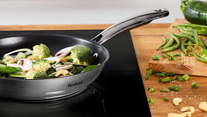 Na obrazie widoczna jest patelnia z logo Tefal, na której znajdują się warzywa, takie jak brokuły i fasolka. Obok na drewnianej desce leżą pokrojone warzywa i orzechy nerkowca.