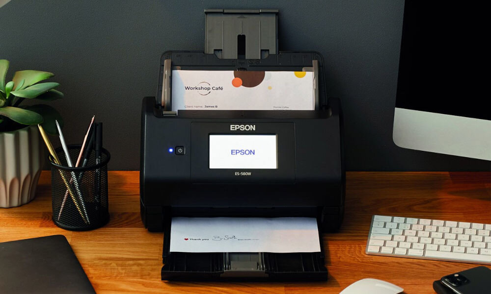 Skaner EPSON WorkForce ES-580W Wi-Fi prezentacja skanera od przodu na biurku wchodząca i wychodząca kartka w tle ciemna ściana obok skanera akcesoria