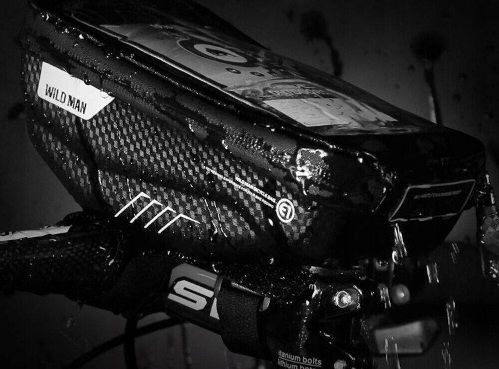 Czarna torba rowerowa na kierownicę z widocznym logo \'WILDMAN\' i wzorem przypominającym włókno węglowe. Torba jest mokra, co sugeruje odporność na wodę.