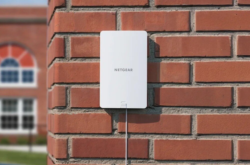 Punkt dostępu NETGEAR WAX610Y Urządzenie z napisem NETGEAR zamontowane pionowo na ścianie z czerwonej cegły z widocznym od dołu białym przewodem biegnącym w dół, opis