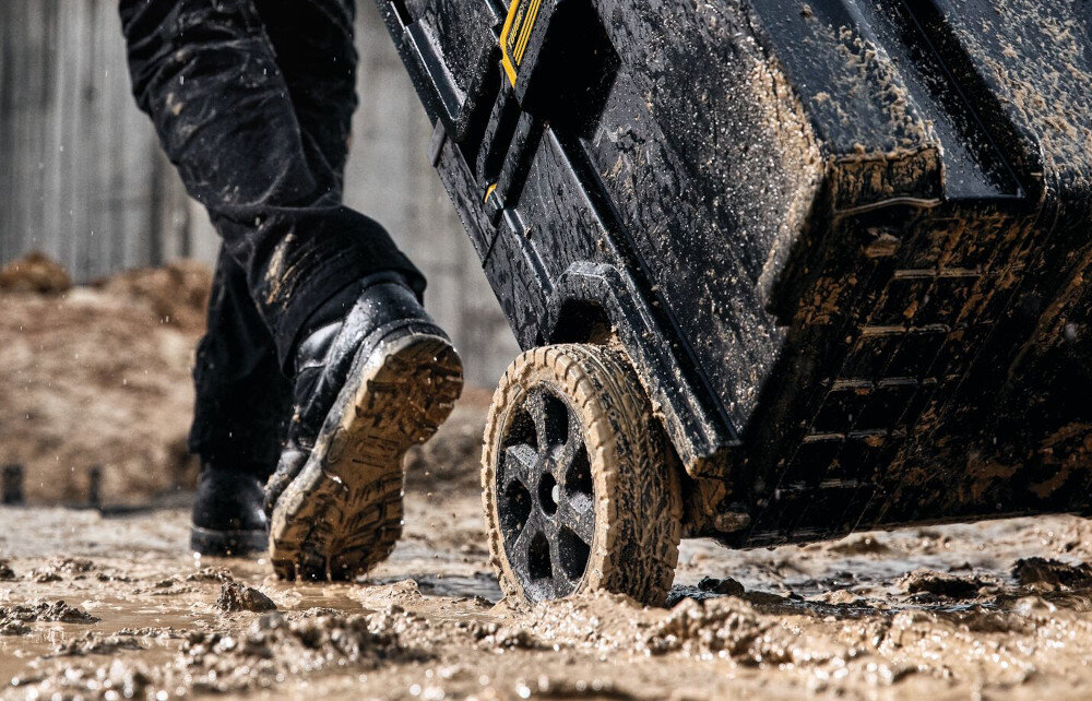 Zestaw skrzynek DEWALT Toughsystem 2.0 DWST83401-1 (3 szt.) plac budowy błoto zbliżenie na kółko osoba ciągnie wózek transportowy po błocie wózek transportowy wytrzymałe 8-calowe koła wysuwany uchwyt swobodne przemieszczanie skrzyń szerokie wzmocnione uchwyty boczne ogumowane uchwyty górne