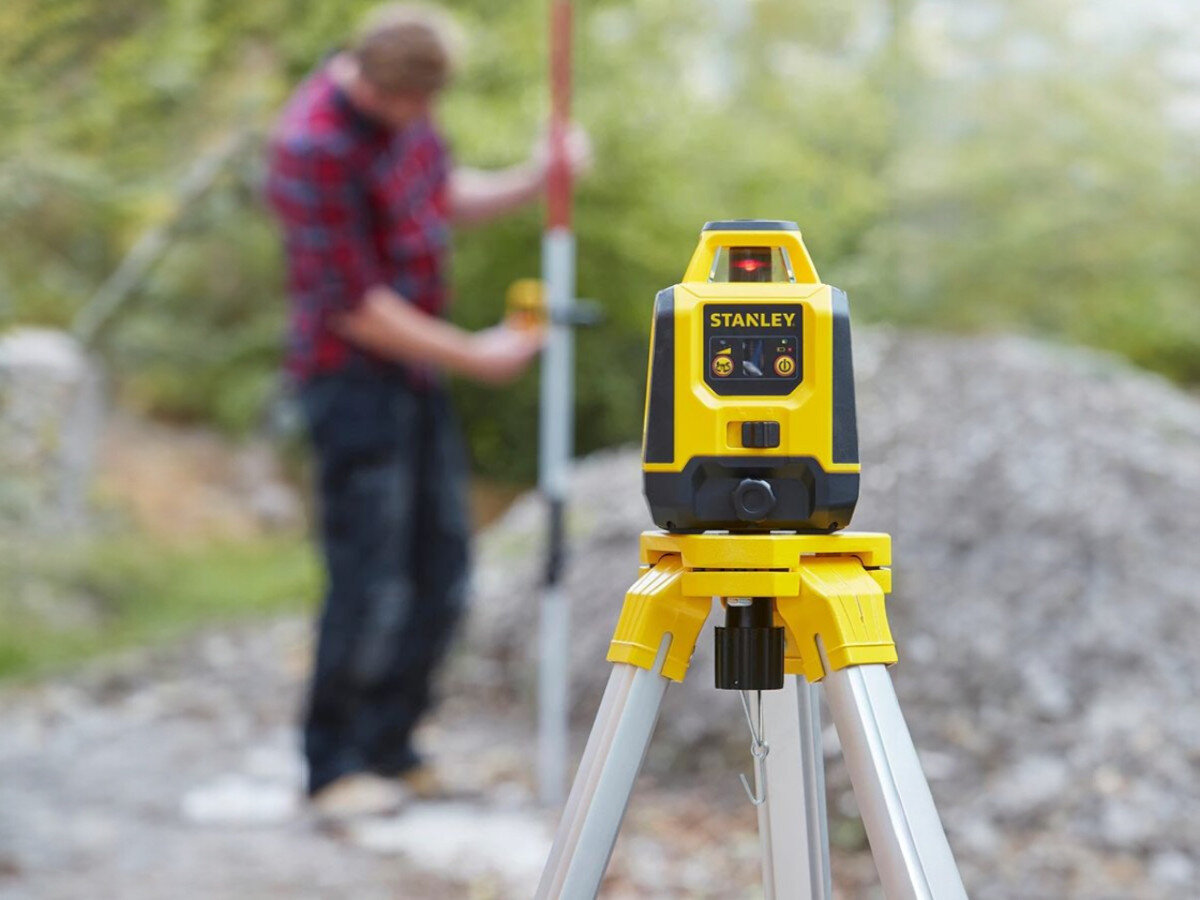 Laser rotacyjny STANLEY STHT77616-0, poziom, rotacja, pomiary, budowa, Niwelator Stanley na statywie w terenie, w tle pracownik z łatą pomiarową.