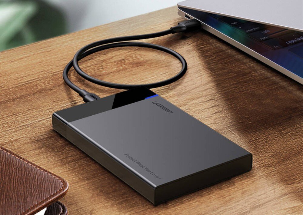 Obudowa dysku UGREEN 50743 Czarna, prostokątna obudowa dysku leżąca na drewnianym blacie, podłączona czarnym kablem USB do srebrnego laptopa, z widocznym napisem 'UGREEN' oraz 'Protect What You Love !' oraz świecącą niebieską diodą