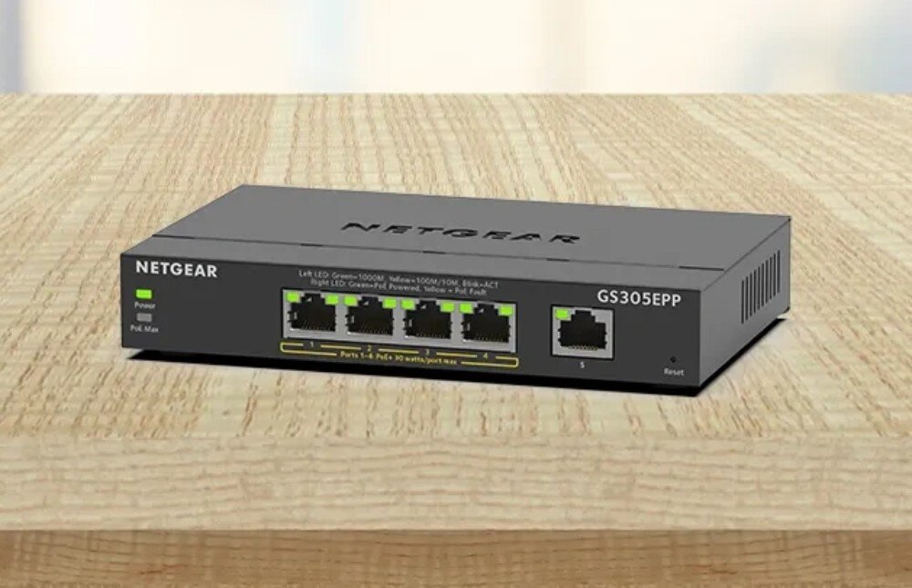 Switch NETGEAR GS305EPP-100PES Przełącznik sieciowy stoi na jasnym, drewnianym stole w oświetlonym pomieszczeniu, opis