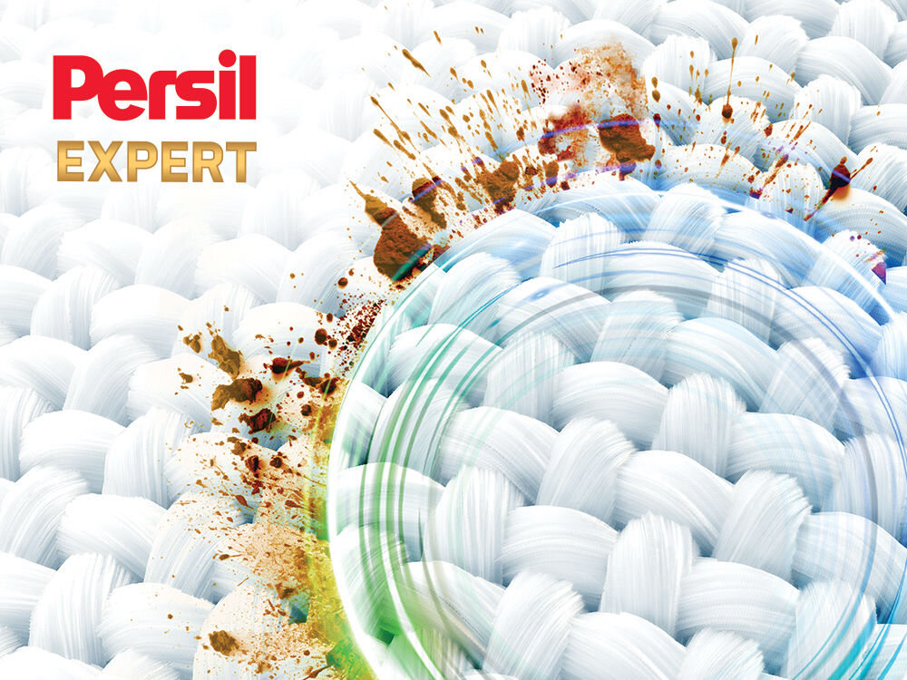 Proszek do prania PERSIL Zdjęcie ilustrujące działanie Persil Expert. Powiększenie struktury tkaniny z widocznymi, rozpryskanymi plamami brudu (błoto, kawa). Część plam jest usuwana przez niewidoczną siłę w kształcie błękitno-zielonego pierścienia, symbolizującego głębokie czyszczenie. Logo: Persil Expert