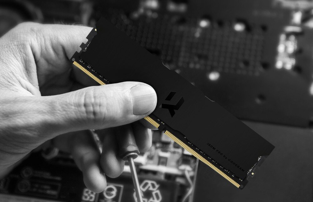 Pamięć RAM GOODRAM IRDM Pro Dłoń trzymająca dwa czarne moduły pamięci RAM typu DIMM z widocznymi złotymi stykami na tle rozmazanego podzespołu komputerowego, jakość, testy