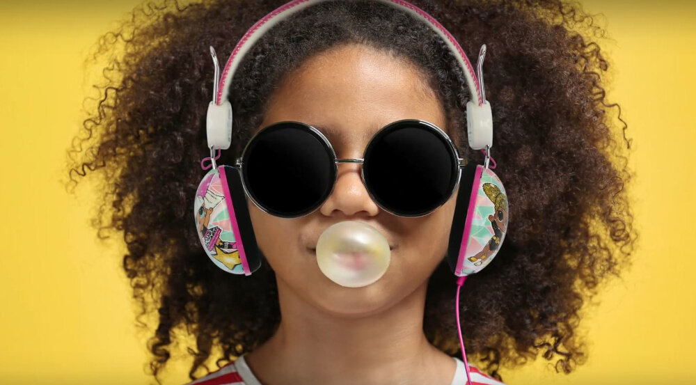 Dziecko nosi czarno-czerwone słuchawki nauszne z motywem Harry\'ego Pottera i okulary przeciwsłoneczne, dmuchając balon z gumy do żucia. Tło jest jednolite, żółte.
