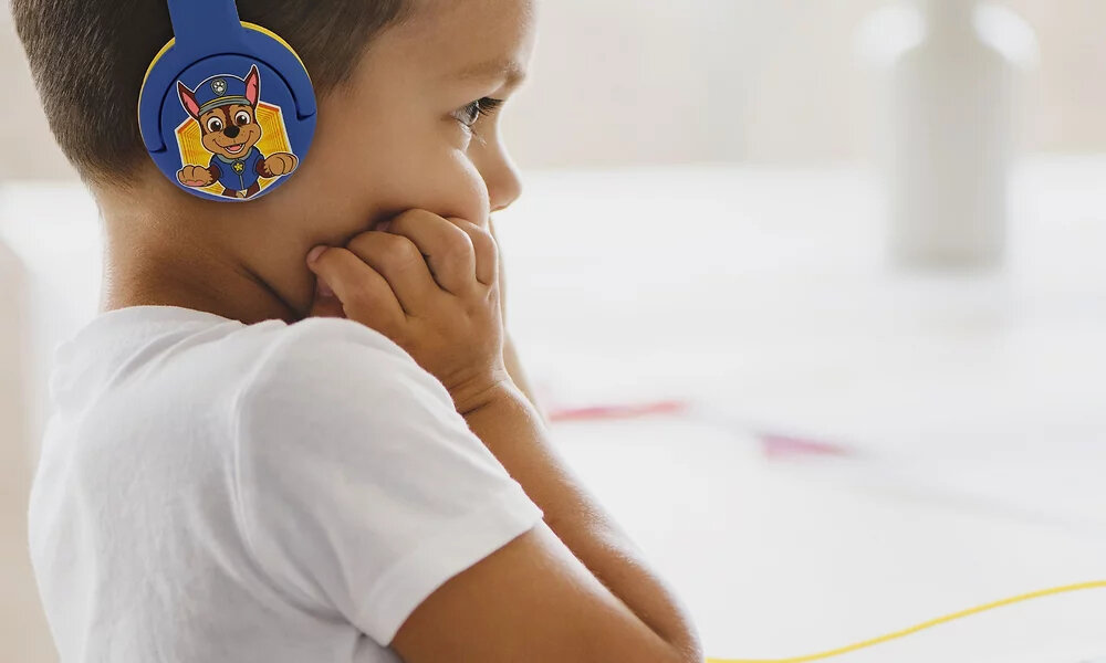 Dziecko nosi niebiesko-żółte słuchawki z postacią z bajki Psi Patrol. W tle widać rozmyty biały element dekoracyjny.