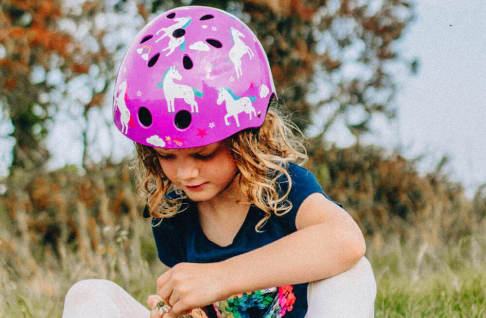 Kask HORNIT Unicorn Fioletowy dla Dzieci (rozmiar S) dziecko w kasku na tle trawnika homologacja CE EN 1078 certyfikat CPSC zgodność z wymaganiami bezpieczeństwa przy prawidłowym założeniu pomaga chronić głowę dziecka przed urazami produkt wspiera codzienną aktywność dziecka w sposób odpowiedzialny i zgodny z normami