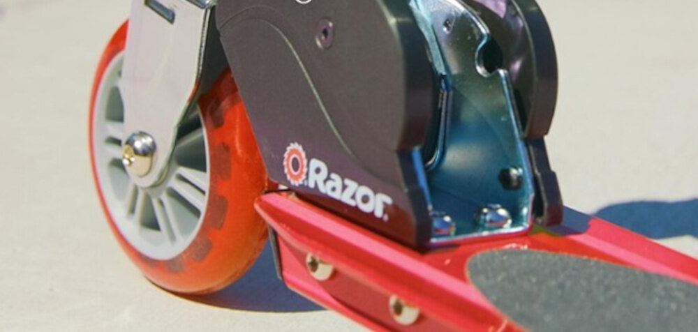 Hulajnoga RAZOR Sport S Czerwony zbliżenie na przednie koło na tle ulicy kauczukowe koła z łożyskami ABEC-5 płynna jazda trwałość średnica 100 mm stabilna jazda łatwe manewrowanie odporne na ścieranie i dobrze tłumią wstrząsy