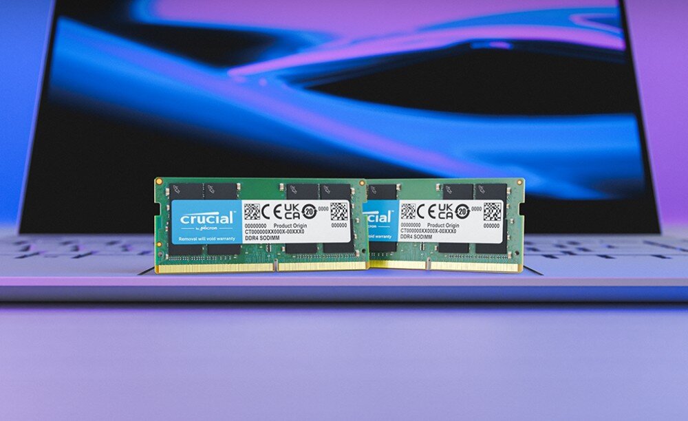 Pamięć RAM CRUCIAL Dwa zielone moduły pamięci ustawione pionowo przed otwartym laptopem z niebieskim ekranem w tle, jakość