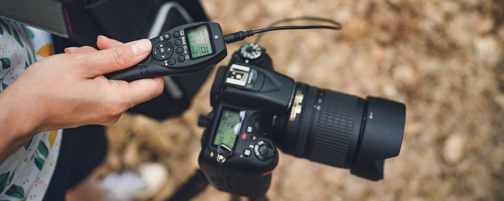 Osoba trzyma pilot bezprzewodowy z wyświetlaczem i przyciskami, podłączony do aparatu fotograficznego z dużym obiektywem.