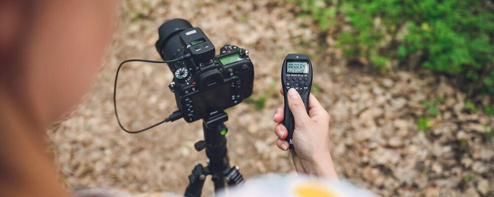 Osoba trzyma pilot bezprzewodowy z wyświetlaczem pokazującym \'00:00:00\'. W tle widoczny jest aparat fotograficzny zamontowany na statywie.