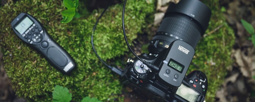 Na obrazie widać aparat fotograficzny z zamontowanym urządzeniem marki Newell oraz pilot bezprzewodowy leżący na mchu. Wyświetlacz na urządzeniu pokazuje \'CH:01\'.