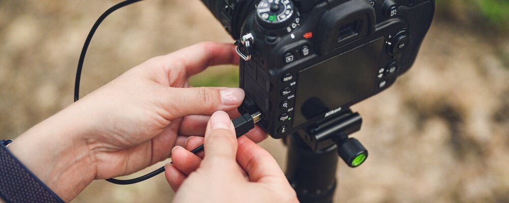 Osoba podłącza kabel do aparatu fotograficznego zamontowanego na statywie. Aparat ma widoczny ekran i kilka przycisków sterujących.