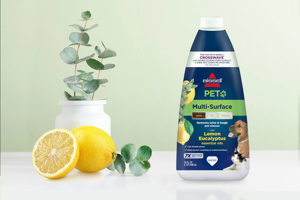 Na obrazie znajduje się butelka płynu do mycia podłóg BISSELL Pet Pro Multiformula 2550 1000 ml z etykietą zawierającą napisy: \'Multi-Surface\', \'Lemon Eucalyptus essential oils\'. Obok butelki widoczne są cytryny i gałązki eukaliptusa w białym wazonie.