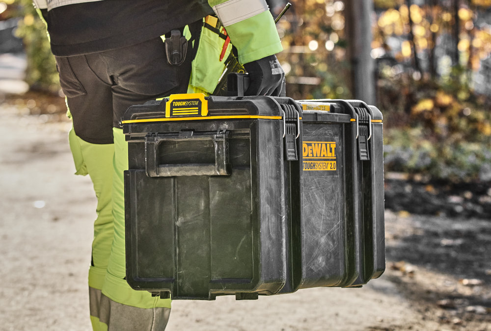 Skrzynka narzędziowa DEWALT DWST83342-1 plac budowy osoba niesie skrzynkę w dłoni obudowa z tworzywa sztucznego odporna na uszkodzenia mechaniczne i działanie czynników zewnętrznych metalowe zatrzaski i metalowe zawiasy udźwig do 50 kg
