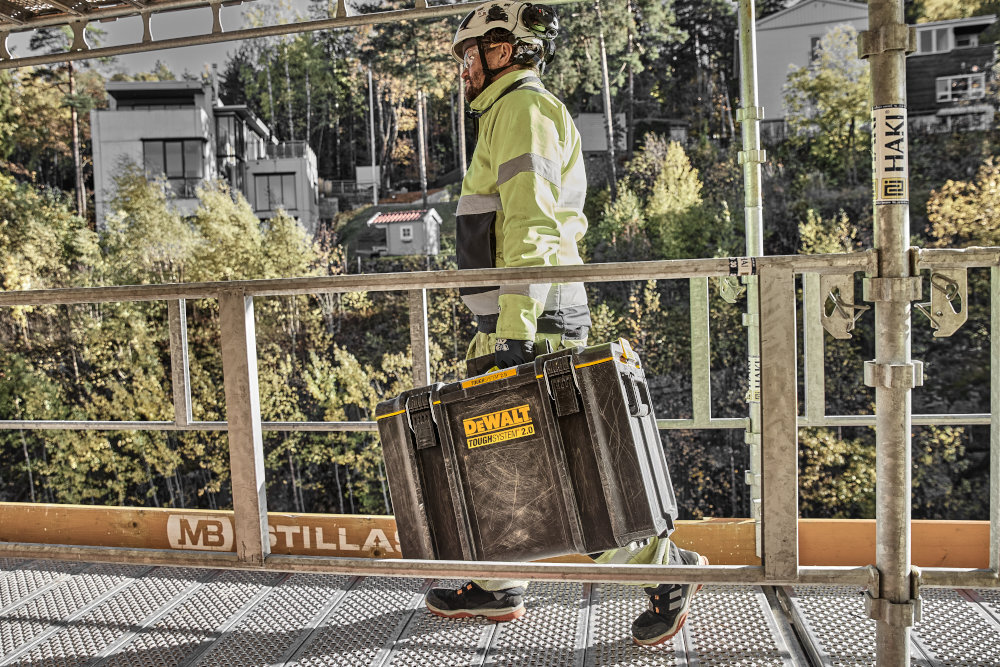 Skrzynka narzędziowa DEWALT DWST83342-1 plac budowy osoba idzie bo metalowym moście w dłoni trzyma skrzynkę w tle kilka budynków i rośliny wyjmowaną tackę na często używane narzędzia skraca czas przygotowania do pracy ułatwia utrzymanie porządku