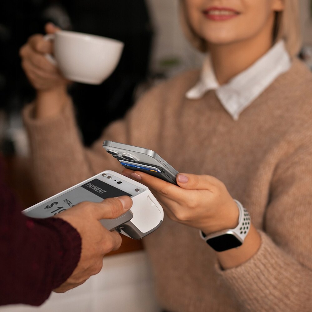 Osoba trzymająca filiżankę kawy płaci telefonem z przymocowanym etui na karty przy terminalu płatniczym. Na ekranie terminala widoczny jest napis \'PAYMENT\'.