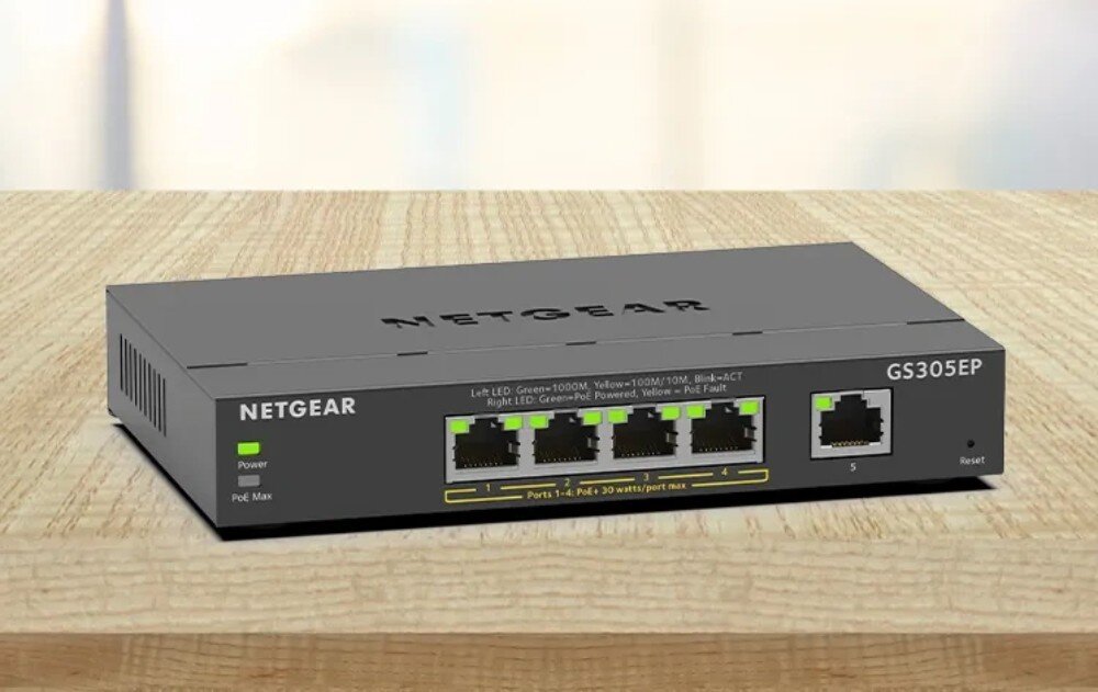 Na drewnianym blacie znajduje się szary switch NETGEAR GS305EP z pięcioma portami Ethernet i wskaźnikami LED. Na urządzeniu widoczne są napisy: \'NETGEAR\', \'GS305EP\', oraz oznaczenia portów i funkcji PoE.