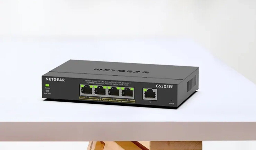 Na białym stole znajduje się czarny switch NETGEAR GS305EP z pięcioma portami Ethernet i oznaczeniami LED. Na obudowie widoczne są napisy: \'NETGEAR\', \'GS305EP\', \'Power\', \'PoE Max\'.