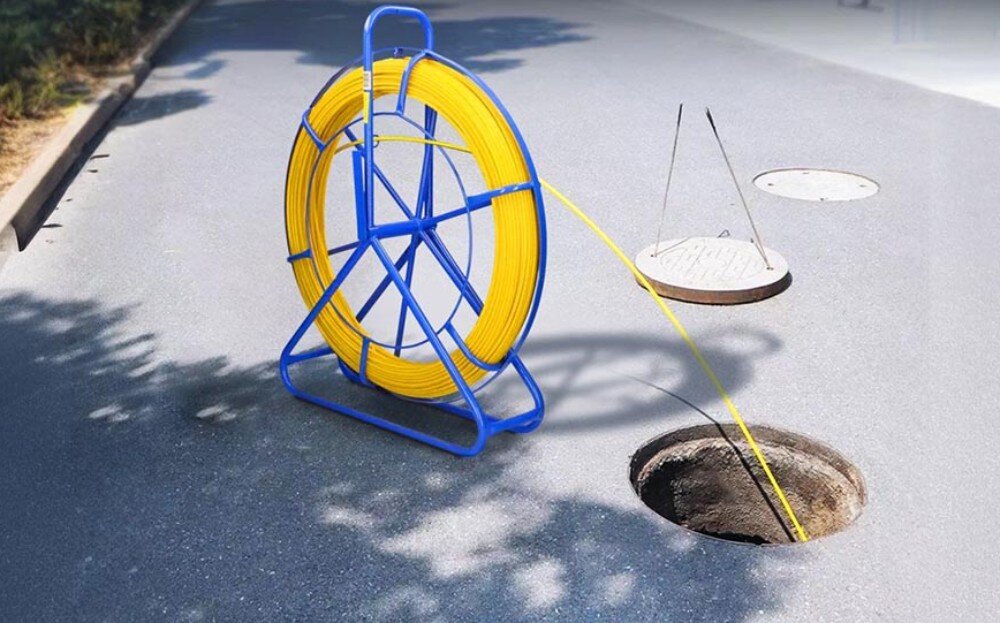 Pilot do przeciągania kabli EXTRALINK EX.16026 Niebieski stojak z żółtym przewodem ustawiony na asfalcie obok otwartego włazu kanalizacyjnego, do którego wprowadzony jest przewód